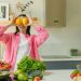 A woman playfully holding lemons over her eyes in a bright, lively kitchen setting filled with fresh vegetables.