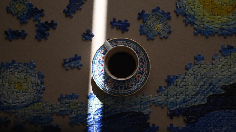 Artistic shot of coffee cup on a Van Gogh puzzle, illuminated by soft light.